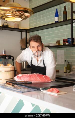 Männlicher Sushi-Koch, der im asiatischen Restaurant auf dem Tisch steht und Rohes Fischfilet auf Schneidebrett legt Stockfoto