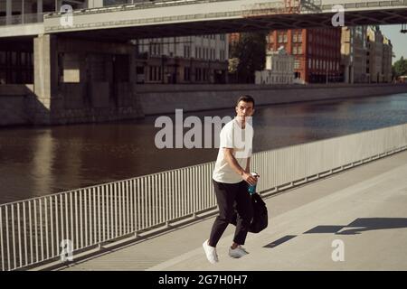 Männlicher Athlet mit Sporttasche, der im Sommer auf dem Damm in der Stadt nach oben geht Stockfoto