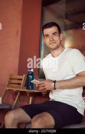 Lächelnder männlicher Athlet, der mit einer Flasche Wasser im Straßencafé sitzt und nach dem Training in der Stadt ein Mobiltelefon benutzt, um die Kamera zu betrachten Stockfoto