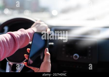 Hand hält das Handy vor dem Hintergrund des Lenkrads des Autos. Abgelenkter Fahrer mit MOBILTELEFON. Telefon mit einem leeren schwarzen Geröll Stockfoto