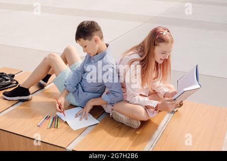 Der Schuljunge und das Schulmädchen lehnen sich auf einer Bank im Schulhof in der Nähe des Schulgebäudes zurück. Studieren mit Büchern und Copybook Stockfoto