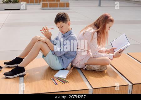 Der Schuljunge und das Schulmädchen sitzen auf der Bank auf dem Schulhof in der Nähe des Schulgebäudes. Studieren mit Büchern und Copybook Stockfoto