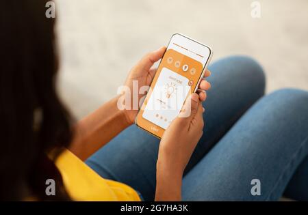 Verwalten der Smart Home-App über das Mobiltelefon zu Hause, Geräte, Lichtverhältnisse, einfache Steuerung. Millennial african american Frau steuert Smartphone mit Lampe Stockfoto