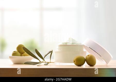 Feuchtigkeitsspendende kosmetische Creme mit Olivenextrakt für die Haut mit Zweig und Oliven auf Holztisch im Zimmer. Vorderansicht. Stockfoto