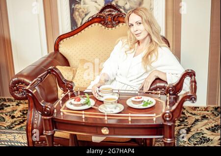 Schöne junge Erwachsene blonde Frau sitzt in einem Stuhl. Auf dem Tisch zwei Tassen und 2 Teller mit Kuchen Stockfoto