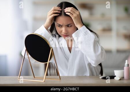 Wütende junge asiatische Dame im weißen Bademantel sitzt im Schlafzimmer, überprüft ihre grauen Haare, Blick auf den Spiegel, enttäuscht Gesichtsausdruck, Kopie spac Stockfoto
