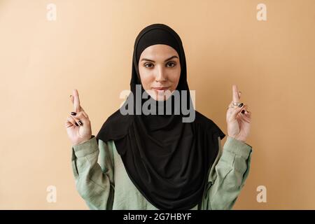 Ein Porträt der positiven muslimischen Frau mit durchbohrter Nase in Hijab, die mit gekreuzten Fingern auf etwas hofft, während sie im orangen Studio steht Stockfoto