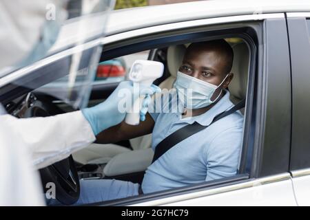 Schwarzer Mann Fahrer trägt eine schützende Gesichtsmaske, sitzt in einem weißen Auto, Arzt in Schutzanzug überprüft afroamerikanischen Kerl Körpertemperatur Stockfoto