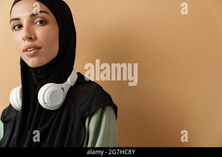 Ein Porträt der muslimischen Frau in schwarzem Hijab mit Kopfhörern, die im orangen Studio steht Stockfoto