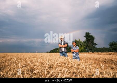 Vater und Sohn stehen nach erfolgreicher Aussaat und Wachstum auf ihrem Weizenfeld. Sie bereiten sich auf die Ernte vor. Regenbogen am Himmel dahinter Stockfoto