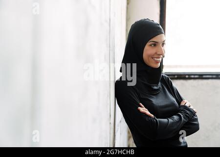 Junge muslimische Sportlerin im Hijab lacht, während sie im Haus steht Stockfoto