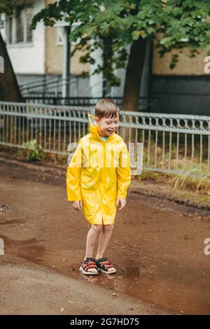 Ein Junge in einem gelben Regenmantel geht im Regen draußen. Ein Kind springt auf die engen Wege. Springt durch schlammige Pfützen. Glückliche und unbeschwerte Kindheit Stockfoto