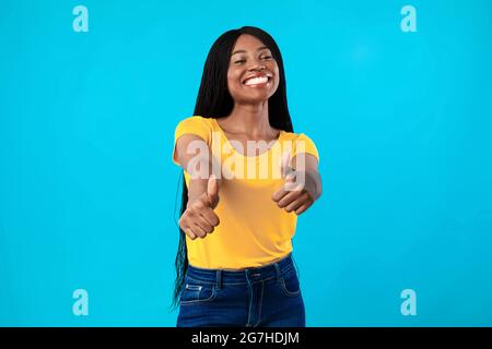 Ich mag es. Toothy Afroamerikanische Frau Gestikuling Thumbs Up Mit Beiden Händen Genehmigung Etwas Posieren Über Blue Studio Hintergrund. Millennial Weiblich Stockfoto