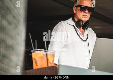 Cooler Discjockey in lässigem weißen Hemd und Sonnenbrille, der für einen Drink in der Nähe der Soundbox draußen während des dj-Sets steht - Disk Jockey oder Barkeeper wo Stockfoto