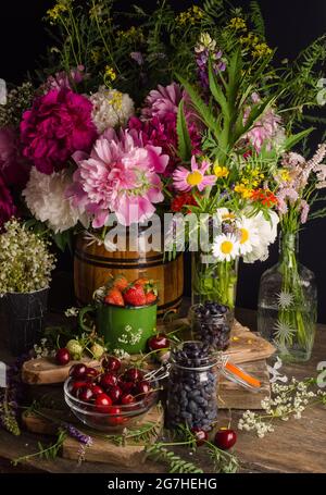 Schickes Sommerstill-Leben mit Pfingstrosen und Erdbeeren, Kirschen und Geißel auf einem alten Holztisch auf schwarzem Hintergrund in einem rustikalen Stall Stockfoto