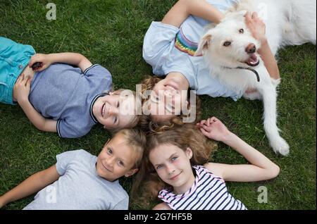 Brüder und Schwestern verschiedener Altersgruppen liegen auf dem Gras und schauen nach oben. Fröhliche, freundliche, fröhliche Jungen und Mädchen mit einem weißen Hund. Die Gesellschaft des Lächelns Stockfoto