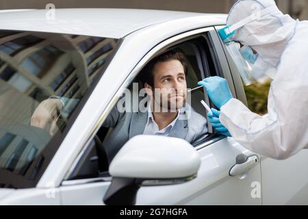 Arzt, der einen Nasenabstrich von einer Person zur Untersuchung auf eine mögliche Covid-19-Infektion einnahm. Sanitäter in Hazmat-Anzug macht Test mit Millennial stattlichen Fahrer in p Stockfoto