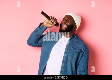 Schöner schwarzer Mann in Beanie und Jeanshemd, der Karaoke singt, Mikrofon hält und auf rosa Hintergrund steht Stockfoto