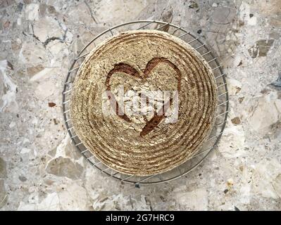 Grundansicht eines runden Sauerteig-Laibs Brot, das in der Kruste herzförmig geritzt ist und auf einem Drahtgestell auf einer Marmorarbeitsfläche abkühlt. Stockfoto