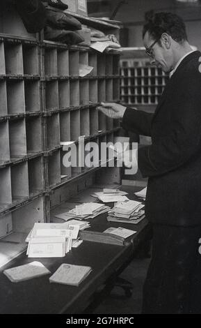 In den 1950er Jahren, historisch, sortierte ein männlicher GPO-Arbeiter Briefe oder Post in den Kubby oder Taubenlöcher auf einem Holzregal für die verschiedenen Straßen des Bezirks, London, England, Großbritannien. Stockfoto