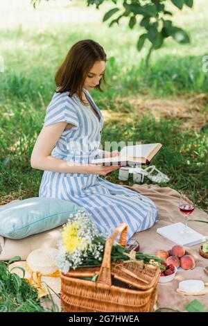 Hübsche Frau, die während des Picknicks ein Buch neben Wein, Obst und Korb auf der Decke liest Stockfoto