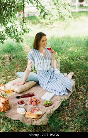 Junge Frau hält ein Glas Wein in der Nähe von Essen, Buch und Korb auf einer Decke im Park Stockfoto