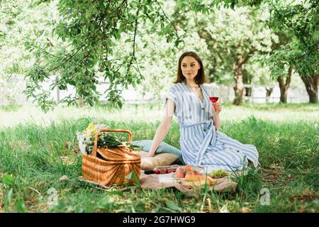 Hübsche Frau hält Wein in der Nähe von Früchten und Blumen im Park Stockfoto