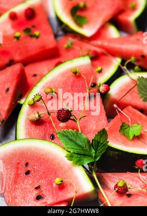 Auf einem grauen Tisch liegen rote Scheiben köstlicher saftiger Wassermelone mit wilden Erdbeeren. Stockfoto