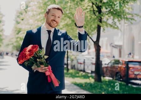 Fröhlicher, männlicher Geschäftsmann mit Blumenstrauß, der jemanden im Stadtpark begrüßt Stockfoto