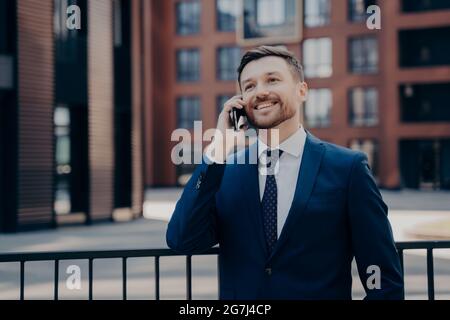 Ein männlicher Büroangestellter genießt angenehme Telefongespräche, während er neben dem Bürogebäude im Freien steht Stockfoto