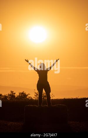 Hintergrundbeleuchtetes Foto der Silhouette einer Frau, die während der goldenen Stunde auf einem Strohballen mit ausgestreckten Armen und der Sonne hinter ihr thront. R Stockfoto