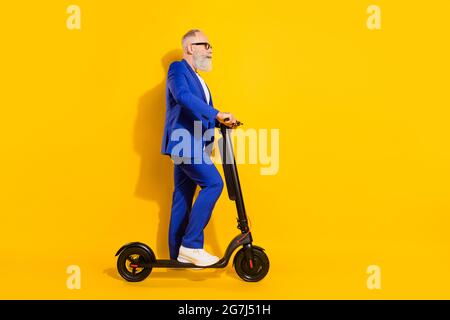 Ganzkörper-Profil Foto von grauem Bart alten Mann Fahrt Roller tragen Brille blauen Anzug isoliert auf gelbem Hintergrund Stockfoto