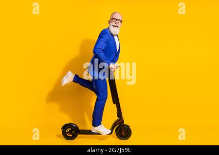 Ganzkörper-Profil-Foto von lustigen grauen Bart im Alter Mann Fahrt Roller tragen Brille blauen Anzug isoliert auf gelbem Hintergrund Stockfoto