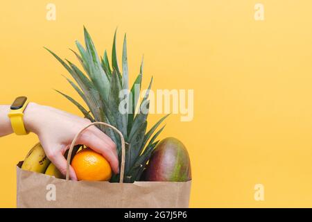Einkaufstasche aus Papier, gefüllt mit tropischen Früchten, mit Frauenhand auf gelbem Hintergrund. Speicherplatz kopieren. Stockfoto