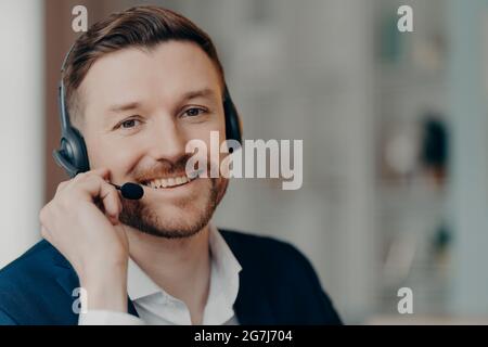 Lächelnder, erfahrener Geschäftsmann mit guter Bartstimmung trägt ein Headset in formeller Kleidung, das vor verschwommenem Hintergrund posiert. Remote-Job Stockfoto