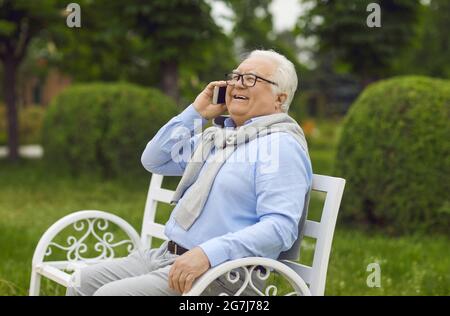 Glücklicher älterer Mann, der auf dem Handy spricht, während er auf der Bank im grünen Sommerpark sitzt Stockfoto