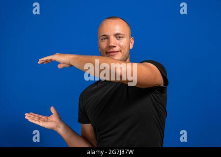 Junger Mann mit schwarzem Hemd auf isoliertem blauem Hintergrund, der einen imaginären Copyspace auf der Handfläche hält, um eine Anzeige einzufügen. Imagination-Konzept Stockfoto