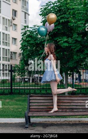 Eine Ballerina in einem Korsett-Kleid geht auf Zehenspitzen auf einer Bank im Park und hält Ballons in den Händen Stockfoto