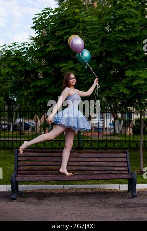 Eine lächelnde, langbeinige junge Frau in einem blauen kurzen Korsett-Kleid geht barfuß auf einer Bank und hält fliegende Ballons Stockfoto