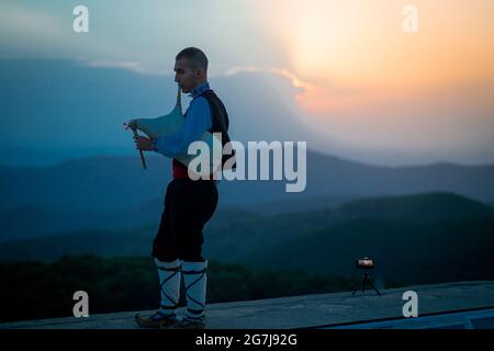 Ein junger Dudelsackläufer in Volkstracht spielt auf einem Berggipfel, wenn die Sonne aufgeht, am 01. Juli 2021, Shipka, Bulgarien Stockfoto