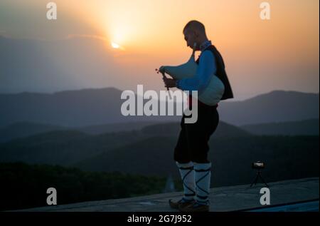 Ein junger Dudelsackläufer in Volkstracht spielt auf einem Berggipfel, wenn die Sonne aufgeht, am 01. Juli 2021, Shipka, Bulgarien Stockfoto