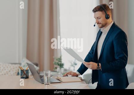 Serious man excutive Arbeiter gibt Präsentation des Geschäftsplans hält Papier Dokument steht in der Nähe des Desktops gekleidet formell verwendet modernen Laptop und Stockfoto