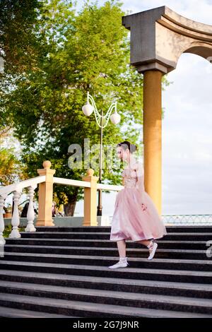 Eine junge Ballerina in einem rosa Seidenkleid läuft im Sommer in einem Stadtpark anmutig die Stufen in einem Bogen mit Säulen am Flussufer entlang Stockfoto