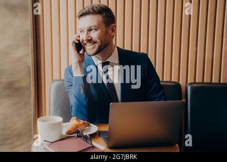 Lächelnder, unrasierte, gutaussehender Mann im formellen Anzug, der im Café per Handy telefoniert Stockfoto
