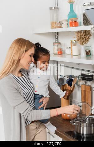 Glückliche Frau hält adoptiert und überrascht afroamerikanischen Kind beim Blick auf Kochtopf Stockfoto