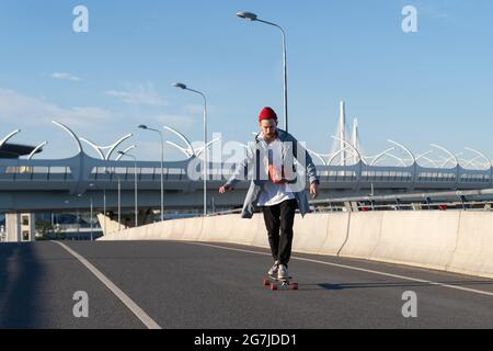 Junger Mann fährt Longboard auf der Brücke. Sorgloser Hipster-Mann im trendigen Fashion-Outfit auf Skateboard Stockfoto