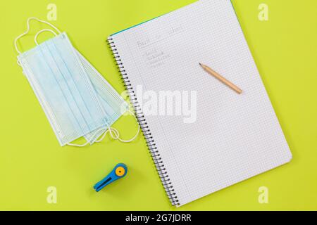 Auf einem hellgrünen Hintergrund befinden sich zwei OP-Masken, ein Locher und ein Notizbuch mit einer Reihe von Schulmaterialien, die mit Bleistift geschrieben sind. Stockfoto