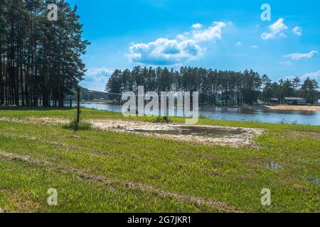 Der Volleyballplatz im Freien liegt neben dem See und auf der anderen Seite befindet sich ein Strand. Bäume und blauer Himmel im Hintergrund. Stockfoto