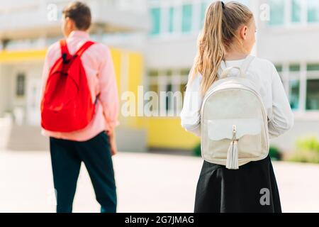 Zwei Kinder mit Rucksäcken gehen zur Schule, Junge und Mädchen Schüler, Kinder mit Schultaschen über den Schultern auf dem Hintergrund der Schule, Retu Stockfoto