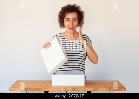 Eine junge Frau mit lockigen Haaren öffnet eine weiße Schuhbox, die sie in einem Online-Shop bestellt hat. Das Unternehmen liefert Pakete schnell zu Ihnen nach Hause. Anniversa Stockfoto